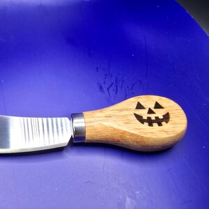 May include: A stainless steel cheese knife with a wooden handle. The handle features a carved jack-o'-lantern face, perfect for Halloween. The knife has a curved blade and a metal ferrule.