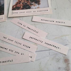May include: A collection of beige paper strips with inspirational quotes printed in black typewriter font. The quotes include "wrap your soul in sunshine", "adventure awaits", "life is beautiful", "perfectly imperfect", "let your light shine", and "thankful".