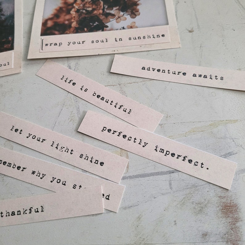 May include: A collection of beige paper strips with inspirational quotes printed in black typewriter font. The quotes include "wrap your soul in sunshine", "adventure awaits", "life is beautiful", "perfectly imperfect", "let your light shine", and "thankful".