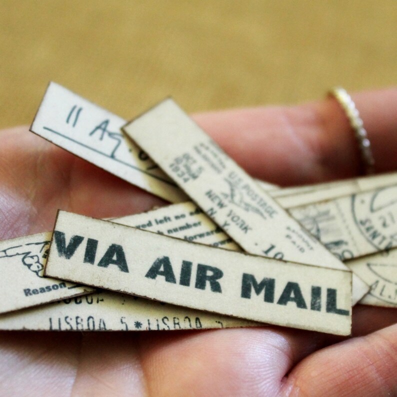 K&ouml;nnte beinhalten: Eine Sammlung von Papieretiketten im Vintage-Stil mit gedrucktem Text und handschriftlichen Notizen. Die Etiketten zeigen S&auml;tze wie "VIA AIR MAIL" und "US POSTAGE NEW YORK". Die Etiketten sind beige mit schwarzer Tinte.