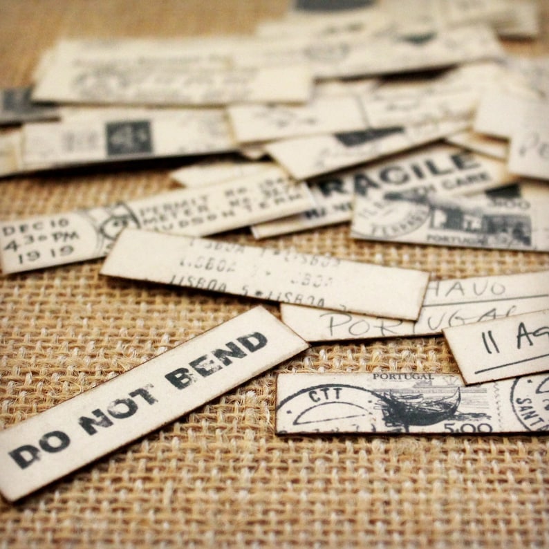K&ouml;nnte beinhalten: Eine Sammlung von Vintage-Papier-Ephemera, darunter rechteckige Etiketten und Aufkleber mit verblasstem schwarzem Text und Stempeln. Ein Etikett zeigt "DO NOT BEND" in fetten Buchstaben. Die Artikel liegen auf einer strukturierten, beigefarbenen Jutefl&auml;che.