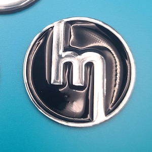 May include: A shiny, circular emblem with a black background and a silver "hm" logo. The emblem is set against a bright blue background, creating a striking contrast. The logo appears to be raised, giving it a three-dimensional effect.