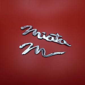 May include: Two chrome-colored car emblems on a red background. The top emblem spells out "Miata" in a stylized font, and the bottom emblem is the letter "M".