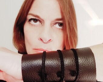 May include: Dark brown leather wrist cuff with three black suede straps. The cuff has a textured surface and is worn on the wrist. The cuff is a fashion accessory.