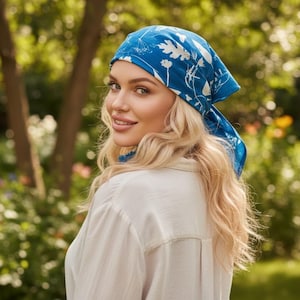May include: A blue headscarf with a white floral design. The scarf is tied around the head. The person is wearing a white button-down shirt. The background is a blurred view of a garden with trees and flowers.