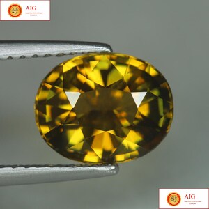 Puede incluir: Una gema ovalada y facetada, sujeta con unas pinzas. La gema es de un color amarillo-naranja vibrante, con múltiples facetas que reflejan la luz. La gema se encuentra sobre un fondo gris neutro. La imagen incluye el texto "AIG" en las esquinas superior izquierda e inferior derecha.