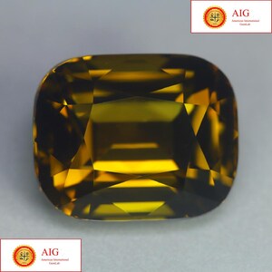 May include: A cushion-cut gemstone with a deep olive-green and golden-brown hue, featuring a faceted surface that creates geometric light reflections. The stone displays a transparent quality with sharp edges and a polished, reflective finish.