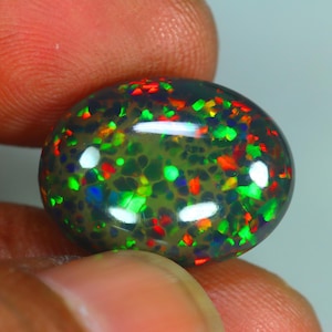 Pode incluir: Uma pedra preciosa de opala negra de formato oval com uma cor base escura e flashes multicoloridos vibrantes de vermelho, verde e azul em toda a superfície. A pedra tem um acabamento polido em corte cabochão com reflexos de luz.