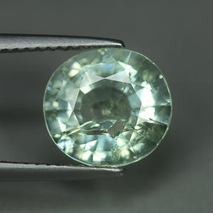 May include: A round, faceted gemstone with a pale green hue. The gem is held by a pair of silver tweezers, showcasing its clarity and cut. The gemstone's facets reflect light, creating a sparkling effect. The overall theme is jewelry and gemstones.