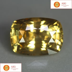 May include: A cushion-cut yellow gemstone with a faceted surface that reflects light, creating bright highlights and deep golden tones. The stone is presented with the AIG American International GemLab logo visible in the corners of the frame.