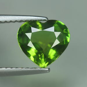 May include: A heart-shaped, faceted gemstone in a vibrant green hue. The gem is held by a silver tweezer, showcasing its intricate cut and brilliant facets. The gemstone's shape and color suggest it could be used in jewelry.