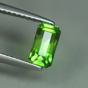May include: A vibrant, rectangular-cut, green gemstone is held by a pair of silver tweezers. The gem has a faceted surface that reflects light, showcasing its clarity and color. The gemstone's bright green hue is the focal point against a soft gray background.