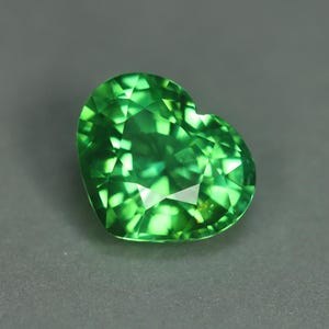 May include: A heart-shaped, faceted gemstone in a vibrant green hue. The gem's surface reflects light, showcasing its intricate cuts and internal brilliance. The gemstone is set against a gray background.
