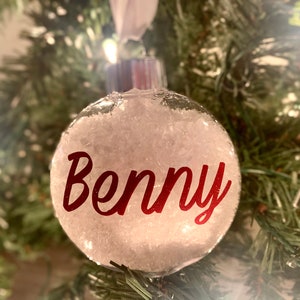 Op de afbeelding: Een heldere, ronde kerstbal gevuld met wit materiaal, mogelijk sneeuw. De naam "Benny" is in rood schrift op de voorkant gedrukt. De bal hangt aan een wit lint tegen een achtergrond van groene kerstboomtakken.