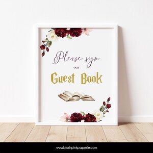 May include: White sign with a burgundy and white floral border. The sign says "Please sign our Guest Book" in gold lettering with an open book illustration.