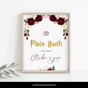 May include: A printable photo booth sign with a burgundy and white floral border. The sign says "Photo Booth" in gold letters with a lightning bolt and "Grab a prop and strike a pose" in cursive writing.
