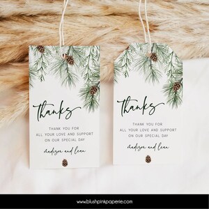 Peut inclure: Deux étiquettes de faveur de mariage blanches avec un motif de branche de pin verte et de cône de pin brun. Le texte sur les étiquettes est "Merci" et "Merci pour tout votre amour et votre soutien pour notre jour spécial" suivi des noms "Madison et Lean".
