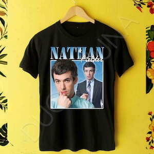 Pode incluir: Camiseta preta com um retângulo branco com uma foto de Nathan Fielder do programa Nathan For You. O texto "Nathan Fielder" está acima da foto.