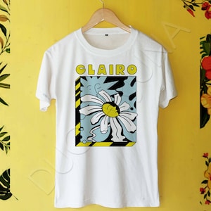 May include: White t-shirt with a graphic of a daisy flower in a yellow and black frame. The text "CLAIRE" is printed above the graphic.