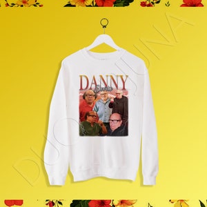 Może przedstawiać: Biała bluza z nadrukiem graficznym obsady serialu telewizyjnego "Zawsze słonecznie w Filadelfii" z tekstem "Danny Devito" w stylu retro.