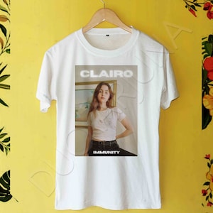 May include: White t-shirt with a photo of a woman in front of a painting. The text "CLAIRE" and "IMMUNITY" are printed on the shirt.
