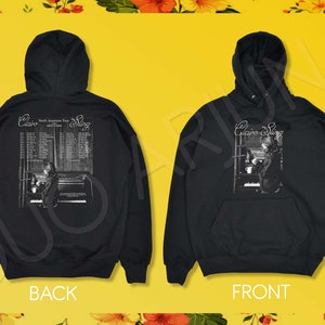 May include: Black hooded sweatshirt with a white graphic print of a person playing a keyboard with the text "Clairo Sling" and "North American Tour Dates" on the back.