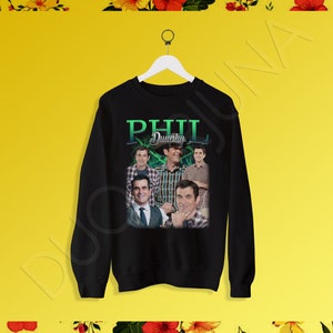 Könnte beinhalten: Schwarzer Pullover mit einem Grafikdruck von Phil Dunphy aus der Fernsehserie Modern Family. Die Grafik zeigt eine Collage von Phils Gesicht in verschiedenen Posen, mit dem Text "Phil Dunphy" in Grün und Weiß.