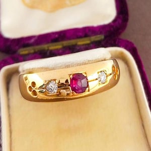 May include: A gold band ring featuring a central round-cut red ruby flanked by two round-cut clear diamonds. The metal surface displays decorative cut-out patterns on both sides of the stones, creating a detailed and polished appearance.