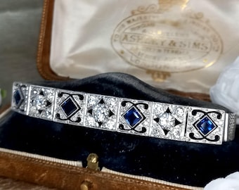 May include: A vintage bracelet with alternating square and round gemstone settings. The bracelet features clear and blue gemstones set in a silver-toned metal. The bracelet is displayed in a velvet-lined jewelry box.