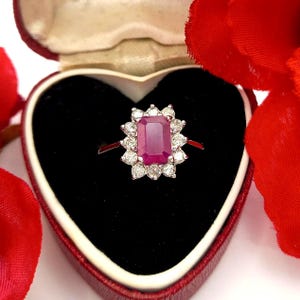 May include: A ruby and diamond ring presented in a heart-shaped box. The ring has a rectangular, deep pink ruby centre stone, encircled by a halo of small, round diamonds. The ring is set on a thin, silver band. The box is red with a black interior.