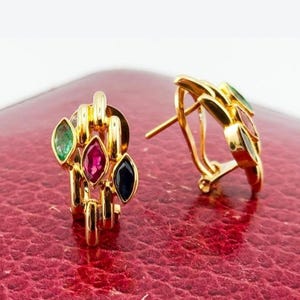 May include: A pair of gold earrings featuring a textured, open-link design. Each earring is set with three marquise-cut gemstones in green, red, and dark blue, arranged vertically within the gold metal structure, secured with a post-back mechanism.