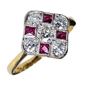 May include: A gold ring with a square face, set with diamonds and rubies. The central diamond is surrounded by four smaller diamonds and four square-cut rubies. The setting is detailed with a silver-colored metal.