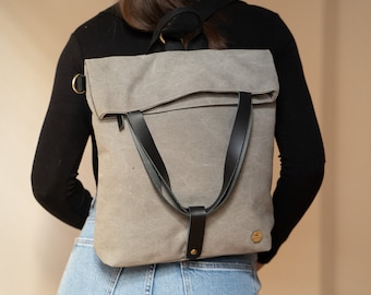 May include: A gray canvas backpack with black leather straps and a gold logo. The backpack has a flap closure and a zippered pocket on the front.
