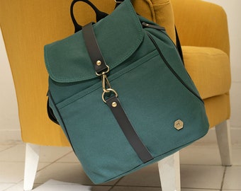 May include: A dark green canvas backpack featuring a fold-over flap with a black leather-like strap and a gold-toned metal clasp. The bag includes black shoulder straps, a top handle, and a small hexagonal gold-toned metal logo badge on the front.