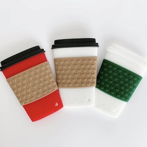 May include: Three coffee cup-shaped silicone accessories in red, white, and white. Each cup has a black lid and a heart-patterned sleeve in tan or green. The cups are designed to resemble to-go coffee cups.