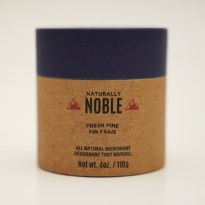 May include: A brown cardboard tube with a blue lid. The label reads "Naturally Noble" with a red mountain logo on either side. The text "Fresh Pine Pin Frais" is printed below the logo. The label also includes the text "All Natural Deodorant Deodorant Tout Naturel" and "Net wt. 4oz./110g".