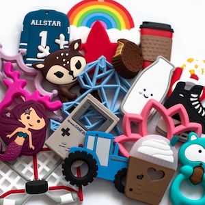 May include: A collection of colourful silicone teethers featuring various designs, including a hockey net, a reindeer, a mermaid, a gameboy, a tractor, a rainbow, a coffee cup, a milk carton, a snowflake, a lotus flower, a popcorn bucket, a hockey skate, a cupcake, and a raccoon.
