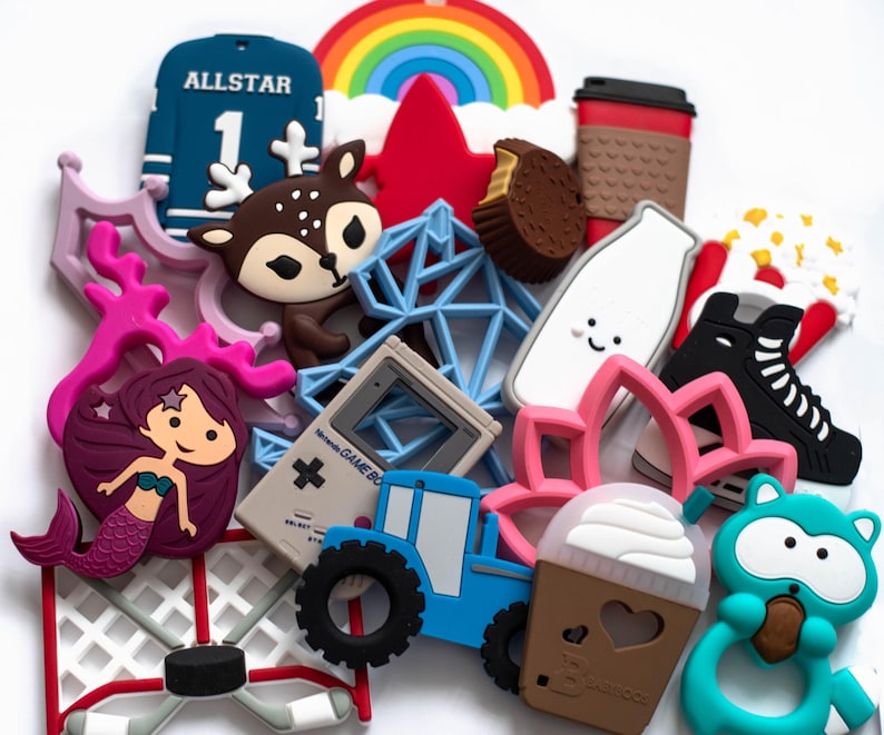 May include: A collection of colourful silicone teethers featuring various designs, including a hockey net, a reindeer, a mermaid, a gameboy, a tractor, a rainbow, a coffee cup, a milk carton, a snowflake, a lotus flower, a popcorn bucket, a hockey skate, a cupcake, and a raccoon.