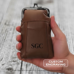 May include: A brown leather coin purse with a metal clasp, held in a hand. The purse has a pocket on the front and the initials "SGC" are engraved on the front. The text "CUSTOM ENGRAVING" is in a white rectangle.