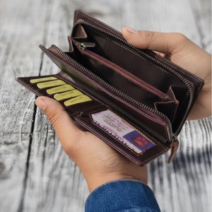 May include: Open brown leather wallet with multiple compartments, including card slots and a zippered section. The wallet is held open, showcasing its interior and a driver's license. The zipper and stitching are visible.