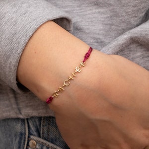 May include: A gold bracelet with the word "CACHE" spelled out in individual letters. The bracelet is on a red string.