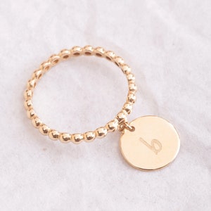 Op de afbeelding: Een gouden ring met kralen en een ronde bedel met de letter "b" erop gegraveerd.