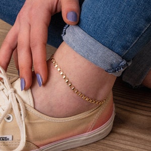 May include: A gold chain anklet with a delicate, linked design. The anklet is worn on a person's ankle, with the foot resting on a light brown wooden surface.