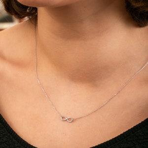Könnte beinhalten: Eine silberne Halskette mit einem Unendlichkeitszeichen auf einer silbernen Kette. Die Halskette wird von einer Person mit einem schwarzen Oberteil getragen.