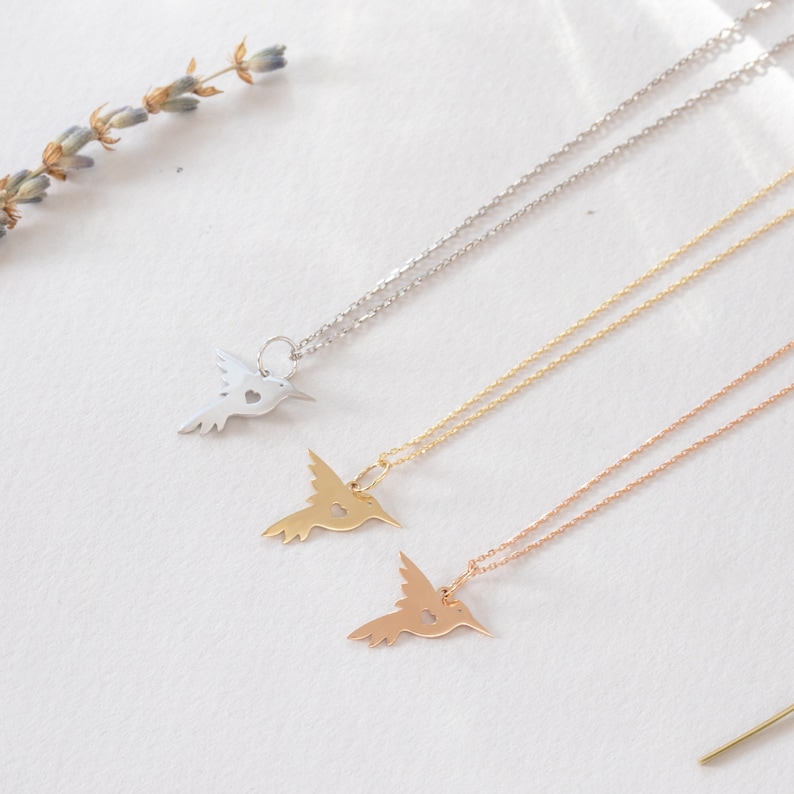 K&ouml;nnte beinhalten: Drei zarte Halsketten mit Kolibri-Anh&auml;ngern. Jeder Anh&auml;nger hat ein ausgeschnittenes Herzdesign. Die Halsketten sind in Silber, Gold und Ros&eacute;gold dargestellt.