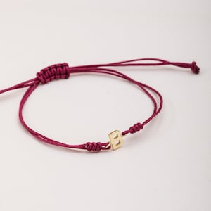Könnte beinhalten: Ein burgunderrotes Armband aus Schnur mit einem goldenen 'B'-Anhänger.