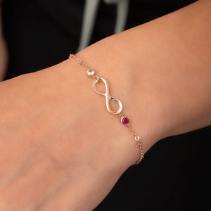Könnte beinhalten: Ein goldfarbenes Armband mit einem Unendlichkeitszeichen, einem kleinen rosafarbenen Edelstein und einem kleineren gelben Edelstein. Das Armband befindet sich am Handgelenk einer Person.