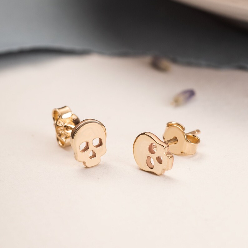 A pair of gold skull stud earrings. Each earring features a small, detailed skull design with eye and nose openings. The earrings are displayed on a white surface, showcasing their polished finish.