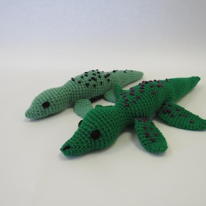 May include: Two crocheted green lizard toys. One lizard is a solid green color, while the other has purple spots. Both lizards have black eyes.
