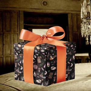 May include: A black gift box with an orange ribbon is wrapped in Halloween-themed paper with a variety of spooky designs. The box is sitting on a white tablecloth in front of a crystal chandelier.